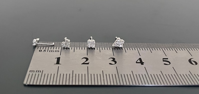 May include: Four silver nose studs with small diamond-like accents are lined up on a ruler, measuring 0.5 mm to 6 mm.