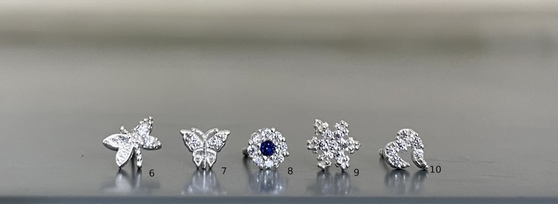 May include: A set of ten silver nose rings with different designs, including a butterfly, a flower, and a snowflake. Each ring is adorned with sparkling cubic zirconia stones.