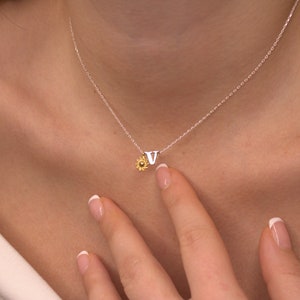 Puede incluir: Un collar de cadena de plata con un pequeño dije de girasol dorado y un dije de plata con la letra 'V'.