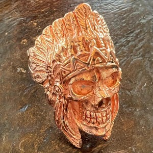 May include: A copper-colored ring featuring a skull design with a Native American headdress. The ring has intricate details, including feathers and geometric patterns, and is set against a dark background.