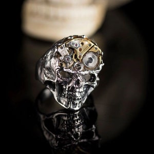 May include: A silver skull ring with a working watch mechanism embedded in the skull. The watch face reads "HOMIS WATCH" and has a silver and gold finish.