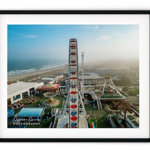 May include: An aerial view of a Ferris wheel with red and white stripes, located at an amusement park by the sea. The Ferris wheel is in the foreground, with the sea and beach in the background.