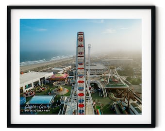 May include: An aerial view of a Ferris wheel with red and white stripes, located at an amusement park by the ocean. The Ferris wheel is in the foreground, with the ocean and beach in the background.