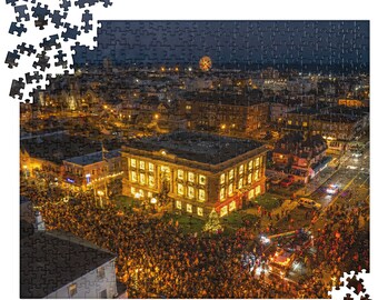 May include: A jigsaw puzzle featuring an aerial night view of a town square illuminated by warm golden lights. A large multi-story building stands centrally, surrounded by a dense crowd of people, a lit Christmas tree, and a distant glowing ferris wheel.