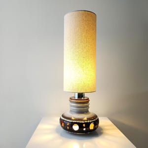 Puede incluir: Una lámpara de mesa de cerámica con una pantalla cilíndrica beige. La base es de cerámica marrón y gris con recortes circulares. La lámpara tiene un cuello cromado y se apoya sobre una superficie blanca. La lámpara está encendida, emitiendo un brillo cálido.