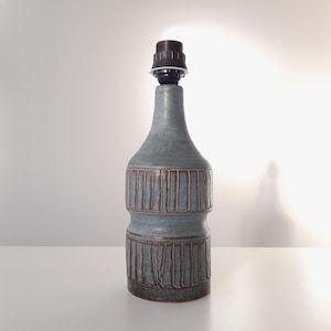 Può includere: Una base per lampada in ceramica in una tonalità grigio-blu tenue, con un design strutturato e a coste in marrone più scuro. La lampada ha una forma a bottiglia con un portalampada nero in alto. Un elemento decorativo per la casa.