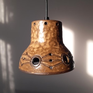Puede incluir: Una lámpara colgante de cerámica marrón con una superficie texturizada y patrones decorativos recortados. La luz cuelga de un cable negro y tiene una base acampanada con aberturas circulares. La lámpara está iluminada por la luz del sol.