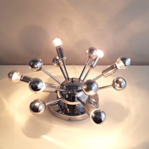 Op de afbeelding: Een plafondlamp in sputnik-stijl met een chromen afwerking, voorzien van een centrale bolvormige naaf met meerdere uitstralende armen. Elke arm eindigt in een gepolijste metalen bol of een lamphouder, wat een stervormig ontwerp creëert.
