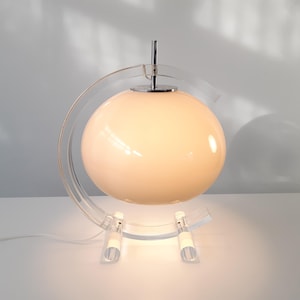 May include: A modern table lamp featuring a spherical, cream-colored glass shade. The lamp is supported by a clear acrylic base and a curved acrylic arm. The lamp has a chrome accent and a power cord.