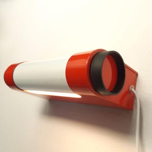 Può includere: Una lampada da parete con corpo cilindrico in metallo arancione, pannello centrale bianco e tappo circolare nero. L'apparecchio emette una luce calda dalla parte inferiore e include un cavo di alimentazione bianco sul lato destro.