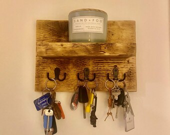 May include: A rustic wooden key holder with a shelf on top. The key holder has three hooks and is mounted on a white wall. A candle jar with the label "SAND + FOG" is on the shelf.