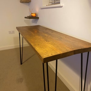 May include: A rustic wooden desk with a natural wood finish and black hairpin legs. The desk is sitting on a beige carpet.