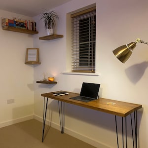 May include: A wooden desk with black hairpin legs and a laptop computer on top. The desk is in a room with white walls and a window with blinds. There is a gold lamp on the wall and a shelf with books on it.
