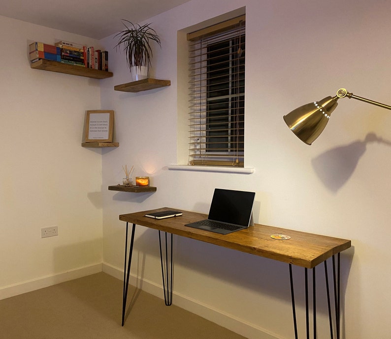 May include: A wooden desk with black hairpin legs and a laptop computer on top. The desk is in a room with white walls and a window with blinds. There is a gold lamp on the wall and a shelf with books on it.
