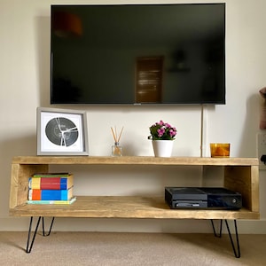 May include: A rustic wooden TV stand with a shelf, featuring hairpin legs and a black gaming console on the lower shelf. The stand is decorated with a framed print, a vase with dried flowers, and a potted plant.
