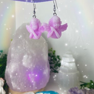 May include: A pair of silver dangle earrings with a light purple angel charm on each earring. The angels are holding their hands together in prayer.