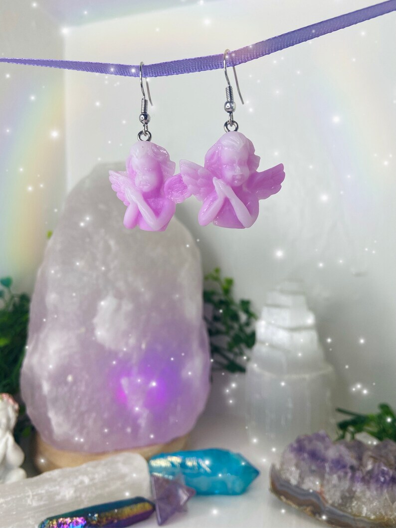 May include: A pair of silver dangle earrings with a light purple angel charm on each earring. The angels are holding their hands together in prayer.
