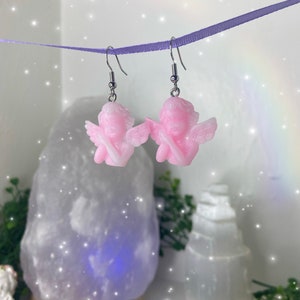 May include: A pair of pink angel dangle earrings with silver hooks. The angels have wings and are holding their hands together in prayer.