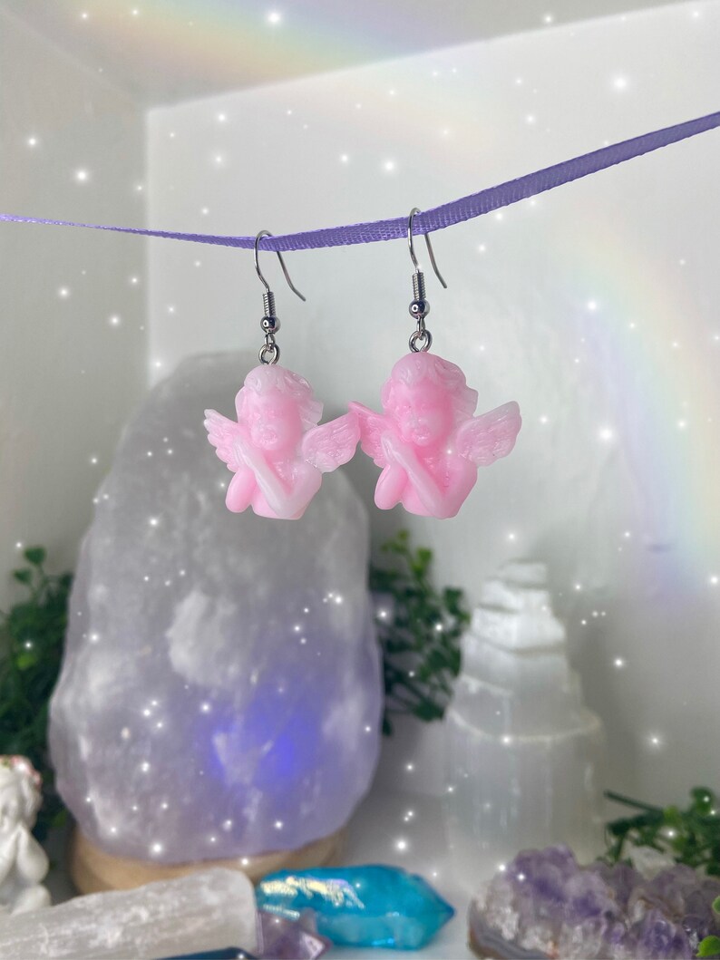 May include: A pair of pink angel dangle earrings with silver hooks. The angels have wings and are holding their hands together in prayer.