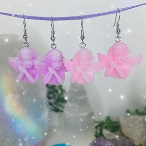 May include: Three pairs of angel-shaped earrings with silver hooks. The angels are made of resin and are available in two colors: light pink and lavender.