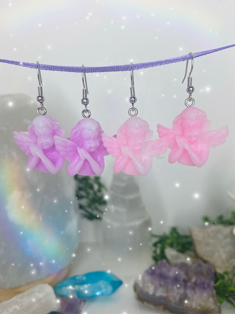 May include: Three pairs of angel-shaped earrings with silver hooks. The angels are made of resin and are available in two colors: light pink and lavender.