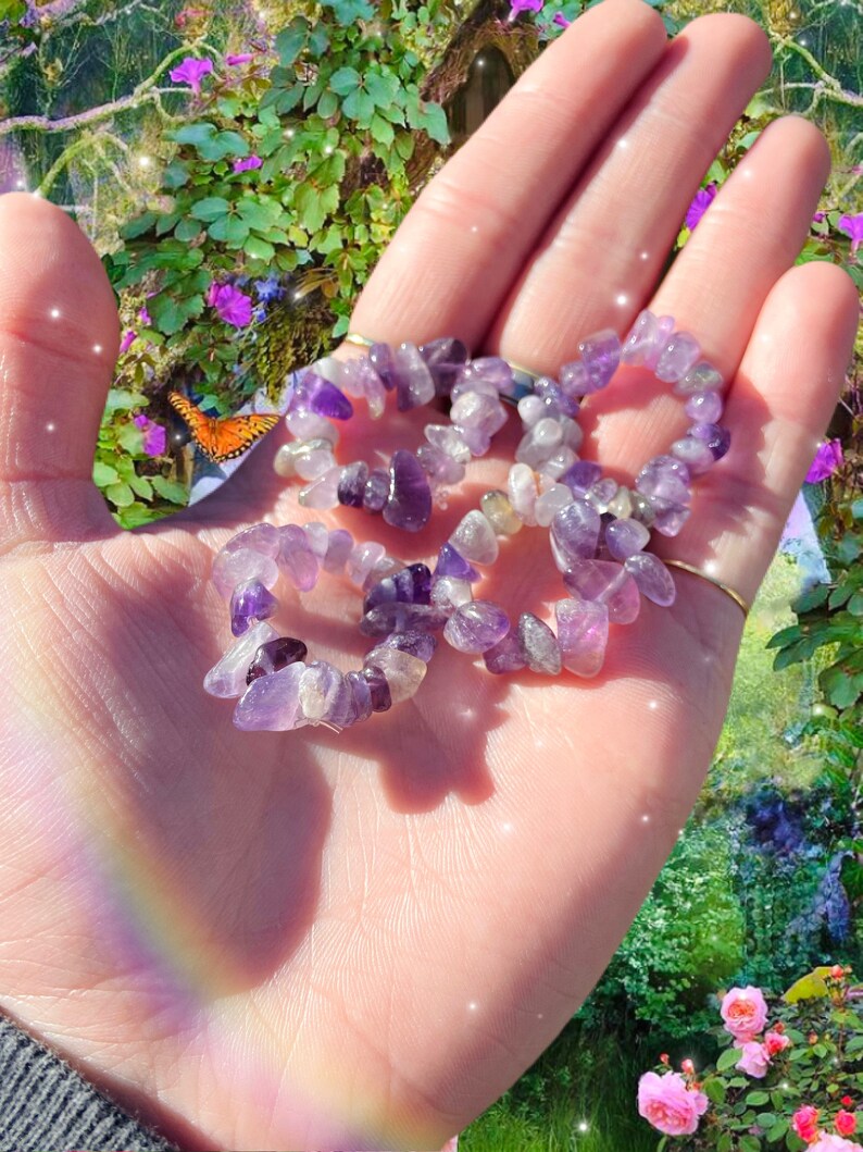May include: A hand holding several amethyst chip stone rings. The rings are made of small, irregularly shaped purple and white stones. The background features a garden scene with flowers, greenery, and a butterfly.