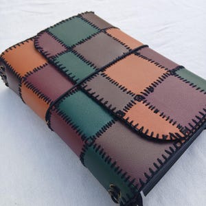 Puede incluir: Un diario rectangular con una cubierta de retazos hecha de paneles verdes, marrones y bronceados. Los paneles están unidos por una costura de látigo negra gruesa a lo largo de los bordes. Un cordón elástico negro rodea el centro para cerrar.