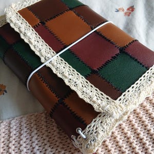 Op de afbeelding: Een rechthoekige journal-omslag met een patchwork-ontwerp van bruine, donkergroene en bordeauxrode vierkanten die met zwarte stiksels zijn verbonden. De omslag is afgewerkt met crèmekleurige kanten randen en vastgezet met een wit elastiek.