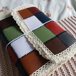 Peut inclure: Une couverture de journal rectangulaire présentant un motif en patchwork de carrés en similicuir dans des tons marron, blanc, bleu marine et vert olive. Les bords sont finis avec une bordure en dentelle crochetée crème et un élastique blanc.