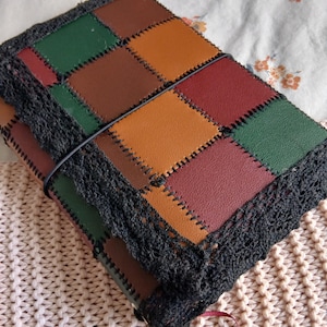 Pode incluir: Um diário retangular com uma capa de patchwork feita de painéis quadrados de aspeto couro em tons de verde, castanho, laranja e bordeaux. As bordas são adornadas com renda de croché preta e um elástico preto mantém a capa fechada.