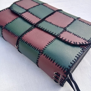 Puede incluir: Un diario rectangular con una cubierta de patchwork de cuadrados alternos en verde oscuro y granate. Los cuadrados están unidos por costuras de festón negras visibles. Un cordón elástico negro envuelve la cubierta y un marcapáginas de cinta negra sobresale por el lateral.