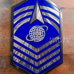May include: A blue and silver metal wall art piece with a shield shape. The design features a blue background with silver stripes and a silver arrow and globe emblem. The text "RUSTY DOG DESIGN CO." is visible.