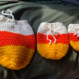 Puede incluir: Tres adornos de maíz dulce de crochet en diferentes tamaños. El adorno más grande es blanco, naranja y amarillo. El adorno mediano es blanco, naranja y amarillo. El adorno más pequeño es blanco, naranja y amarillo.