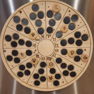 May include: A wooden circular board with a zodiac wheel design. The board has 12 sections, each with a zodiac symbol and a small black circle. There are also small wooden circles with astrological symbols placed on the board.
