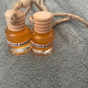 Pode incluir: Dois pequenos frascos de vidro cheios de líquido laranja, cada um com uma tampa de madeira clara e um rótulo branco com o texto "sugared pumpkin" em preto. Os frascos estão ligados por um cordão bege trançado com detalhes dourados.