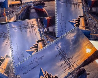 May include: Spiral-bound 2026 calendar featuring a sunset over a harbor with a tall ship and modern architecture. The calendar cover reads "Stu Art Glasgow" and "2026 Calendar" in white script. The image has a blue and orange color scheme.