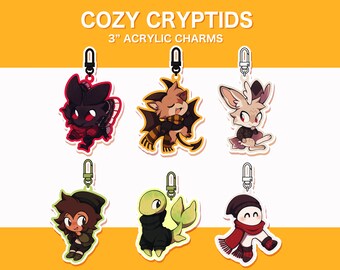 May include: Six 3-inch acrylic charms featuring cute, cartoon cryptids wearing cozy winter accessories. The charms are attached to silver keychains.  The cryptids include a moth, a bat, a deer, a turtle, a lizard, and a snowman.