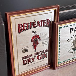 May include: Two framed vintage signs. The first features "BEEFEATER LONDON DISTILLED DRY GIN" with a red-clad figure and the text "London 1873-1924." The second sign reads "PADDY OLD IRISH WHISKEY" with a crest. Both signs have brown frames.