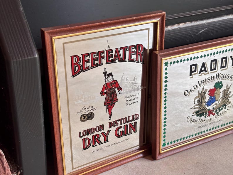 May include: Two framed vintage signs. The first features "BEEFEATER LONDON DISTILLED DRY GIN" with a red-clad figure and the text "London 1873-1924." The second sign reads "PADDY OLD IRISH WHISKEY" with a crest. Both signs have brown frames.