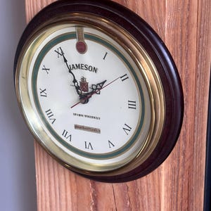 Può includere: Un orologio da parete rotondo con una cornice in legno marrone scuro e una lunetta color oro. Il quadrante è bianco con numeri romani neri e il marchio "Jameson". L'orologio è montato su una superficie di legno.