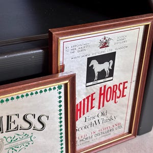May include: Two framed vintage advertisements. One features the text "WHITE HORSE" with a white horse silhouette and "Fine Old Scotch Whisky." The other features the "GUINNESS" logo with a harp. Both have brown frames.