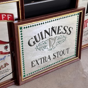May include: A collection of vintage framed bar signs. The central sign features the Guinness logo with the words "EXTRA STOUT". The frames are brown with gold trim. Other signs include "Smirnoff Vodka" and "White Horse" whiskey.