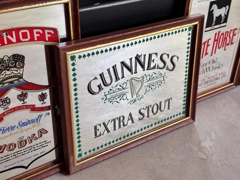 May include: A collection of vintage framed bar signs. The central sign features the Guinness logo with the words "EXTRA STOUT". The frames are brown with gold trim. Other signs include "Smirnoff Vodka" and "White Horse" whiskey.