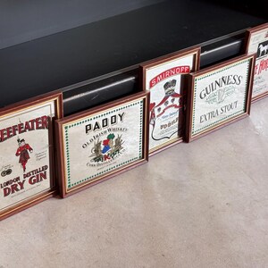 May include: A collection of framed vintage liquor advertisements, including "Beefeater London Distilled Dry Gin," "Paddy Old Irish Whiskey," "Smirnoff Vodka," "Guinness Extra Stout," and "White Horse." The frames are brown, and the ads have a vintage aesthetic.