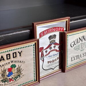May include: A collection of framed vintage liquor signs, including "PADDY Old Irish Whiskey", "Smirnoff Vodka", and "Guinness Extra Stout". Each sign has a brown frame and decorative border.