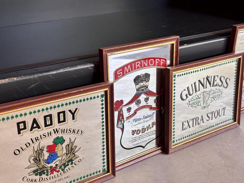 May include: A collection of framed vintage liquor signs, including "PADDY Old Irish Whiskey", "Smirnoff Vodka", and "Guinness Extra Stout". Each sign has a brown frame and decorative border.