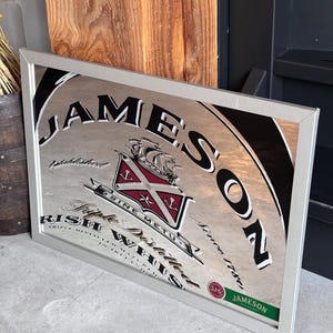 Può includere: Specchio incorniciato con il logo Jameson Irish Whiskey. Lo specchio con cornice argentata presenta il nome del marchio in grandi lettere nere, l'emblema dell'azienda e testo. Lo sfondo è argento riflettente.