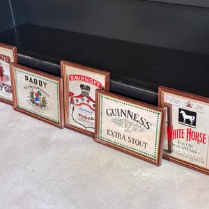 May include: A collection of framed vintage liquor advertisements, including "Beefeater Dry Gin," "Paddy Old Irish Whiskey," "Smirnoff Vodka," "Guinness Extra Stout," and "White Horse Fine Old Scotch Whisky." The frames have a wooden appearance.