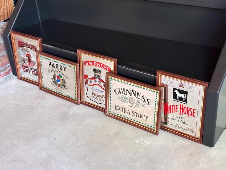 May include: A collection of framed vintage liquor advertisements, including "Beefeater Dry Gin," "Paddy Old Irish Whiskey," "Smirnoff Vodka," "Guinness Extra Stout," and "White Horse Fine Old Scotch Whisky." The frames have a wooden appearance.