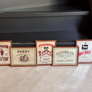 May include: Six framed vintage liquor advertisements, including "Beefeater Dry Gin," "Paddy Old Irish Whiskey," "Smirnoff Vodka," "Guinness Extra Stout," and "White Horse Fine Old Scotch Whisky." Each frame has a brown wooden border.